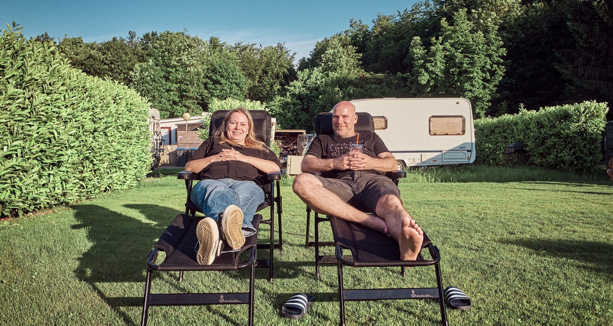 Zwei Personen entspannen sich in Liegestühlen auf dem Camperpark Amsterdam, mit einem Wohnwagen und viel Grün drumherum