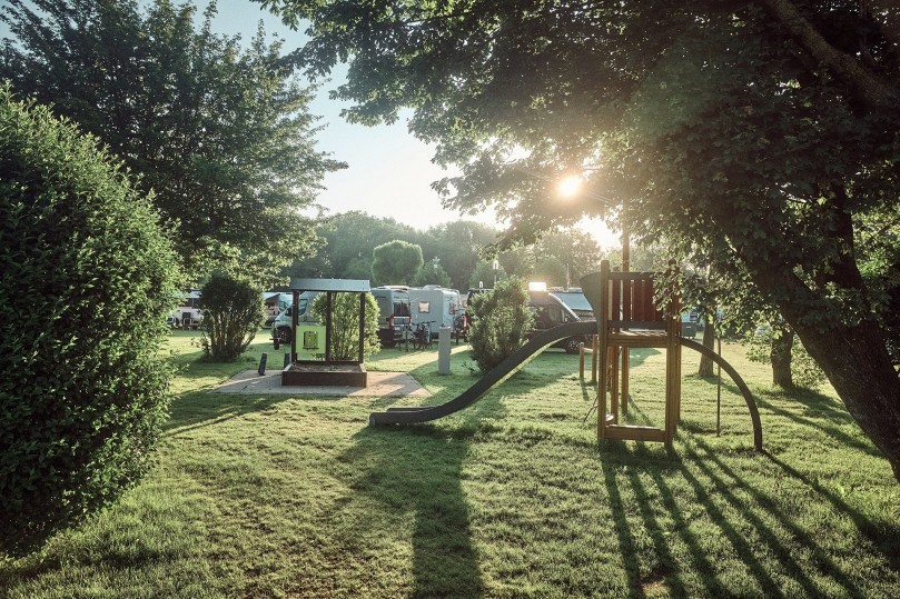 Area giochi e prato del Camperpark Amsterdam, con i campeggiatori sullo sfondo e la luce del sole che attraversa gli alberi
