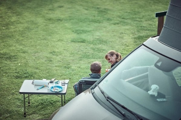 Zwei Personen sitzen entspannt neben einem Wohnmobil auf einer Wiese, mit einem kleinen Campingtisch voller Essen und Getränke